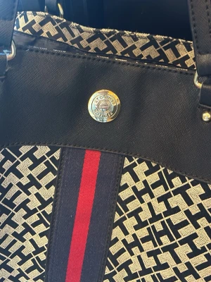Mönstrad shoppingväska med läderdetaljer - Snygg shoppingväska i svart och beige med grafiskt mönster och svarta läderdetaljer. Väskan har dubbla axelband, en dekorativ rand i rött och marinblått samt en rund metallknapp framtill. Perfekt för dig som vill ha en rymlig och trendig väska.