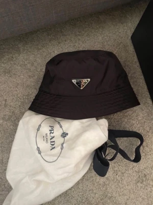 Prada bucket hat - Snygg svart bucket hat från Prada i glansigt nylonmaterial med den ikoniska metalltriangeln framtill. Nytt pris 1500kr MITT ENDAST 550kr kommer utan tillbehör.