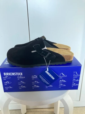Svarta Birkenstock tofflor i mocka - Säljer ett par svarta Birkenstock tofflor i mocka med klassisk slip-in design och justerbart spänne. Fotbädden är i kork och innersulan i mjukt material för extra komfort. Perfekta för chill dagar hemma eller när du vill ha en clean och enkel stil.
