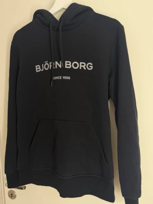 Svart hoodie från Björn Borg - Snygg svart hoodie från Björn Borg med vit logga och texten 'Since 1956' på bröstet. Klassisk känguruficka framtill, justerbar huva med snören.
