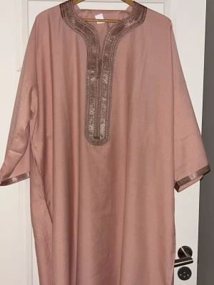 Rosa kaftan / gandoura med broderade detaljer - Säljer en lös och bekväm rosa kaftan med vackra broderade detaljer runt halsringningen och ärmsluten. Plagget har vida ärmar och ett traditionellt snitt, perfekt för dig som gillar unika och stilrena plagg. Helt ny 