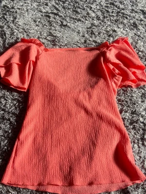 Korallfärgad topp med puffärm - Trendig topp i stark korallfärg med korta puffärmar och fyrkantig ringning. Materialet är lätt strukturerat och ger en snygg textur. Perfekt för dig som vill sticka ut med en färgglad och modern look. Ny och så vacker!