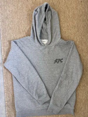 Grå hoodie från APC - Säljer en stilren grå hoodie från APC med svart logga på bröstet. Hoodien har huva, ribbade muddar vid ärmslut och nederkant samt är gjord i mjukt sweatshirtmaterial. Perfekt för en avslappnad och trendig look.