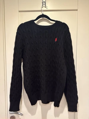 Svart kabelstickad tröja Polo Ralph Lauren - Svart kabelstickad tröja från Polo Ralph Lauren med rund halsringning och röd broderad logga på bröstet. Tröjan har långa ärmar och ribbade muddar vid ärmslut och nederkant. Perfekt för en stilren och klassisk look.