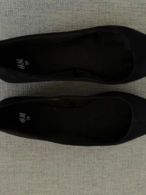 Svarta ballerinaskor från H&M - Klassiska svarta ballerinaskor från H&M i storlek 39. Skorna har rundad tå och är tillverkade i ett mjukt tygmaterial med enkel design. Perfekta att matcha med olika outfits för en stilren look.