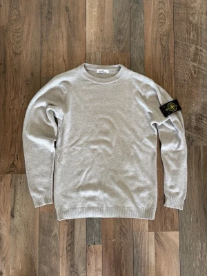 Ljusgrå stickad tröja Stone Island - Snygg ljusgrå stickad tröja från Stone Island med klassisk rund hals och ribbade muddar. Tröjan har den ikoniska Stone Island-loggan på vänster ärm som fästs med knappar. Perfekt för dig som gillar stilrena och trendiga plagg.