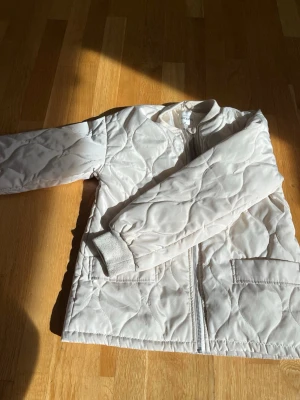 Ljusbeige quiltad vindjacka - Snygg ljusbeige vindjacka med quiltat mönster och raka ärmar. Jackan har dragkedja framtill, två stora fickor och ribbade muddar vid ärmsluten. Perfekt för lager på lager och enkel att matcha med olika outfits.