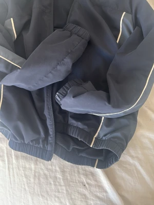 Mörkblå vindjacka med vita detaljer - Snygg mörkblå vindjacka med vita ränder längs ärmarna. Jackan har dragkedja framtill, elastiska muddar vid ärmslut och nederkant samt en sportig look. Perfekt för blåsiga dagar och enkel att matcha med olika outfits. För både 1400kr