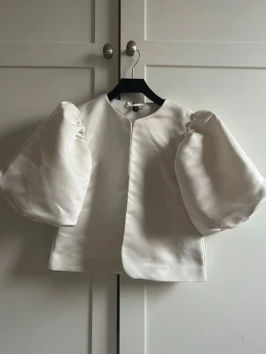 By malina liknanade Vit blus med puffärmar - Stilren vit blus med stora puffärmar och rak passform. Tillverkad i ett glansigt, satinliknande material som ger en lyxig känsla. Perfekt för dig som vill sticka ut med en trendig och elegant look.