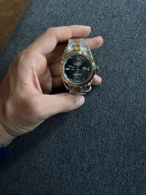 Stilren klocka i guld och silver - Snygg klocka med svart urtavla, guldfärgad räfflad ring och länkarmband i både silver och guld. Klockan har tydliga markeringar och datumvisning. Perfekt accessoar för dig som gillar klassisk men modern stil.