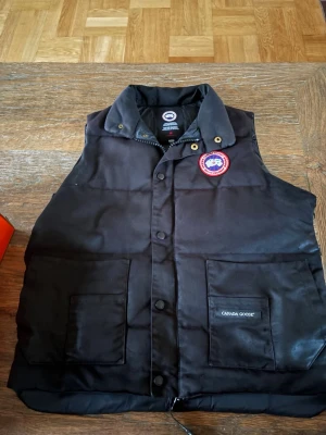Äkta Canada goose väst  - Svart dunväst från Canada Goose med klassisk logga på bröstet och två stora fickor framtill. Västen har hög krage, tryckknappar och dragkedja. Perfekt för lager-på-lager och håller dig varm under kyliga dagar.