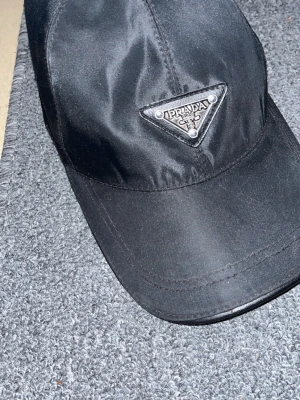 Svart keps från Prada - Snygg svart keps från Prada med det klassiska trekantiga metall-logotypen framtill. Kepsen är tillverkad i ett glansigt material och har justerbart spänne bak för perfekt passform. Perfekt accessoar för en trendig och stilren look.