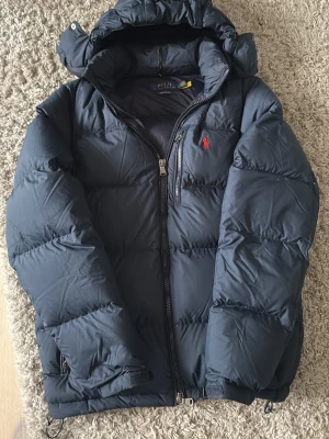 Mörkblå pufferjacka från Polo Ralph Lauren - Snygg mörkblå pufferjacka från Polo Ralph Lauren med huva och dragkedja. Jackan har det klassiska röda Polo-logot på bröstet och är tillverkad i ett tåligt, vadderat material som håller dig varm under kalla dagar. Perfekt för vinter och streetwear-stil. Pris går att diskutera vid snabba affärer. 