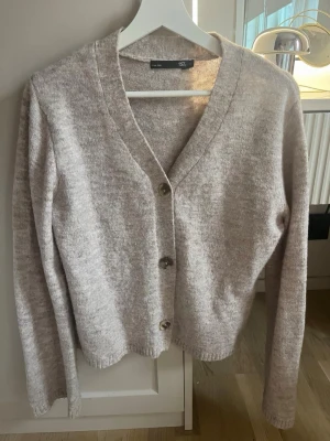 Beige kofta från 157 med knappar - Mysig beige kofta från 157 med v-ringning och tre knappar framtill. Långärmad modell i mjukt stickat material, perfekt att slänga över en t-shirt eller linne. Enkel och stilren design som passar till det mesta.