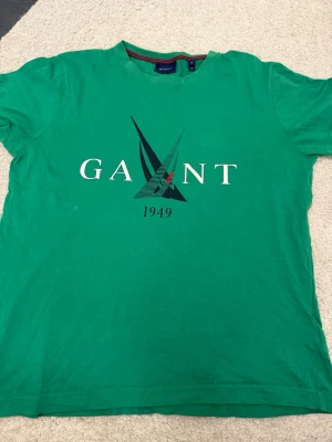 Grön GANT t-shirt med tryck - Snygg grön t-shirt från GANT med stort tryck framtill och texten '1949'. Klassisk passform och rund halsringning. Perfekt till jeans eller shorts för en avslappnad look. Har en liten fläck som syns på bild och går säkert att ta bort med speciella medel 