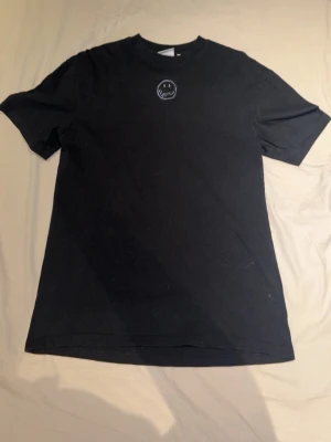 Svart t-shirt med graffiti-tryck - Svart t-shirt med ett litet smiley-tryck på bröstet och stort graffiti-inspirerat tryck på ryggen med texten 'Karl Kani Smiley' och en krona. T-shirten har rund hals och korta ärmar. Perfekt för dig som gillar streetwear och coola detaljer.