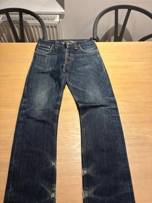 Mörkblå raka nudie jeans med kontrastsömmar - Säljer ett par mörkblå jeans med raka ben och snygga kontrastsömmar. Jeansen har klassisk femficksdesign och detaljerade bakfickor med broderad söm. Materialet är kraftig denim i bomull och passformen är avslappnad men ändå stilren.