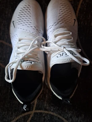 Vita Nike Air Max 270 sneakers - Snygga vita Nike Air Max 270 sneakers med svart logga och detaljer. Ovandelen är i ventilerande mesh och sulan har en tydlig Air-enhet för extra komfort. Skorna har vita skosnören och svart insida, samt dragflik bak för enkel påtagning.