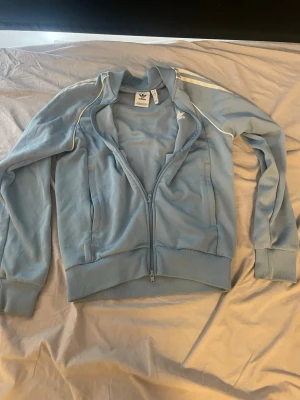 Ljusblå Adidas ziptröja.  (Pris kan diskuteras) - Ljusblå ziptröja från Adidas med vita detaljer och klassiska ränder på axlarna. Tröjan har ståkrage, två fickor framtill och ribbade muddar vid ärmslut och nederkant. Perfekt för en sportig och avslappnad stil.