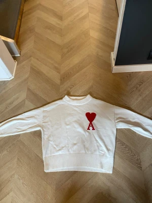 Vit stickad tröja med rött hjärta - Vit stickad tröja med ribbade muddar och bred ribbad nederkant. På bröstet finns ett stort rött hjärta och bokstaven 'A' i samma färg. Tröjan har rund hals och lång ärm, perfekt för en clean och trendig look.