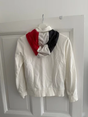 Vit Moncler hoodie - Säljer en vit hoodie från Moncler med dragkedja framtill och två fickor. Huvan har en cool färgblock-design i rött, vitt och svart. Moncler-logga på ärmen. Storlek är 152cm. Nästan aldrig använd, säljer den då den är för liten. 