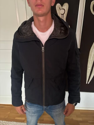 Jacka - Snygg svart vindjacka med dragkedja framtill och stor huva. Jackan har två snedställda fickor med lock och en extra ficka på ärmen. Fodrad insida och justerbar huva med spänne baktill. Perfekt för blåsiga dagar. Jack and Jones premium jacka med storlek M.