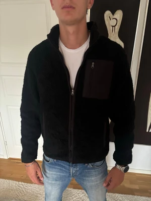 Svart fleece-cardigan med dragkedja - Svart cardiganjacka i mjuk fleece med hel dragkedja framtill och hög krage. Jackan har en bröstficka med dragkedja och två sidofickor. Perfekt för lager-på-lager och chill vardagsstil.