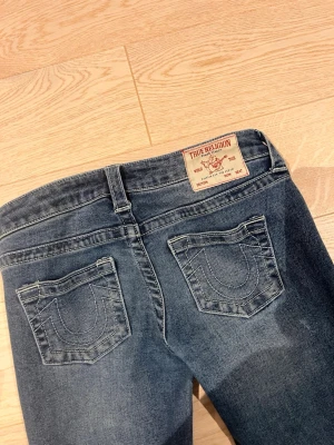 Bootcut jeans från True Religion - Snygga blå bootcut jeans från True Religion med klassiska kontrastsömmar och ikoniska fickdetaljer bak. Jeansen har låg midja och är tillverkade i mjuk denim med lätt tvättad look. Perfekta för dig som gillar en avslappnad men trendig stil.