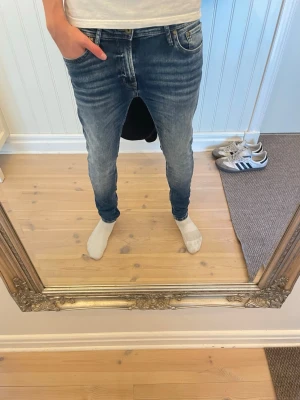 Blå slim fit jeans med tvättade detaljer - Säljer ett par blå slim fit jeans med snygga tvättade detaljer och smal passform. Jeansen har klassisk femficksdesign, normal midja och dragkedjegylf. Perfekta för dig som gillar en modern och avslappnad stil. 29/30