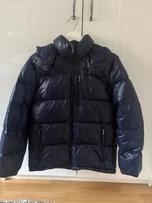 Mörkblå dunjacka från Polo Ralph Lauren - Snygg mörkblå dunjacka från Polo Ralph Lauren med glansig finish och klassisk grön logga på bröstet. Jackan har hög krage, dragkedja framtill, flera fickor med dragkedja och quiltad design. Perfekt för kalla vinterdagar. XL i barn men passar allt från 165-175 beroende på kroppsform. Mycket bra skicka inga defekter 