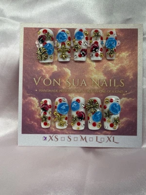 Curiosa (XS);Medium Square - Handgjorda unika press-on naglar med vit bas, dekorerade med blå 3D-blommor, röda och svarta detaljer samt guldiga metalliska dekorationer. Naglarna har en medium lång squareform och är prydda med pärlor och stenar för en unik look.XS: 14 mm, 11 mm, 11,5 mm, 10 mm, 8 mm