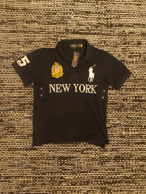 Svart pikétröja Polo Ralph Lauren - Svart pikétröja från Polo Ralph Lauren med vit broderad logga på bröstet, texten 'NEW YORK' och gul broderad detalj. Kortärmad modell med krage och knappslå. Snygg kontrast med vit siffra på ärmen.