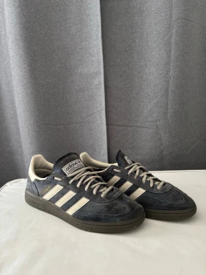 Adidas Spezial sneakers - Säljer ett par Adidas Spezial sneakers i marinblå mocka med klassiska vita ränder på sidorna och vita skosnören. Skorna har en låg profil och mörkbrun sula, samt detaljer i beige och guldtext på sidan. Perfekta för dig som gillar retrostil och streetwear. Kan även passa dig som har storlek 41. Nypris 1250.
