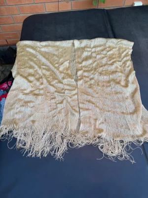 Guldig glittrig sjal med fransar - Säljer en snygg guldig sjal med glittrig finish och långa fransar längs kanten. Sjalen är rektangulär och har en tunn, lätt struktur som ger ett elegant intryck. Perfekt att slänga över axlarna för en extra touch till din outfit.
