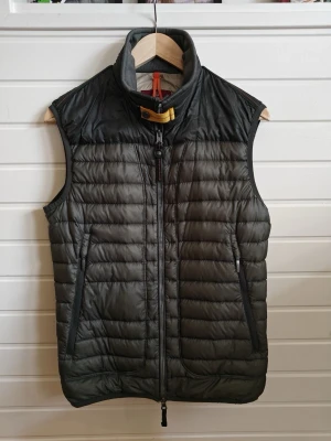 Mörkgrön dunväst med hög krage - Snygg mörkgrön dunväst med hög krage och gul detalj vid halsen. Västen har två dragkedjeförsedda fickor framtill och quiltad design. Perfekt för lager-på-lager och ger en sportig look. Märke syns på ryggen men kan ej identifieras.