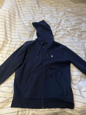 Marinblå hoodie från Polo Ralph Lauren - Snygg marinblå hoodie från Polo Ralph Lauren med dragkedja, huva med dragsko och två fickor framtill. Klassisk liten logga broderad på bröstet. Tillverkad i mjukt material som känns skönt mot huden. Perfekt för en avslappnad och stilren look.