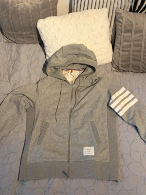 Grå thom browne hoodie - Snygg grå zip-hoodie med huva och dragsko. På vänster ärm finns fyra vita ränder som ger en sportig vibe. Två fickor framtill och en liten vit patch nertill. Tillverkad i mjukt sweatshirtmaterial, perfekt för chill dagar.