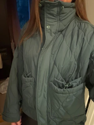Grå pufferjacka med hög krage - Säljer en grå pufferjacka med hög krage och dragkedja samt tryckknappar framtill. Jackan har två stora fickor med dragsko och ett vågigt quiltat mönster. Perfekt för kalla vinterdagar och riktigt mysig att bära.