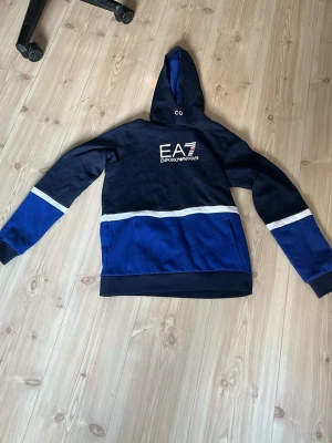 Blå EA7 Emporio Armani hoodie - Snygg hoodie från EA7 Emporio Armani i marinblå och koboltblå färg med vita detaljer. Stor logga på bröstet och huva med dragsko. Tillverkad i mjukt material, perfekt för en avslappnad och trendig look.