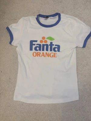 Vit Fanta Orange t-shirt med blå detaljer - Säljer en vit t-shirt med blå krage och ärmslut. Framsidan har ett stort Fanta Orange-tryck i blått och orange med grönt blad. T-shirten har en klassisk passform och är gjord i mjukt bomullsmaterial. Perfekt för dig som gillar retro och coola tryck!
