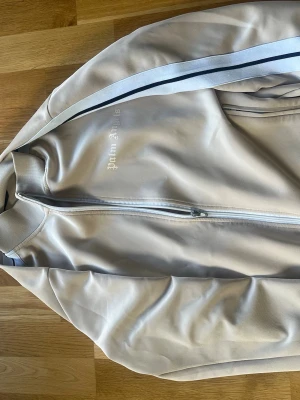 Palm angels biege tröja - Snygg beige palm angels ziptröja med hög krage och dragkedja framtill. Tröjan har vita och mörkblå ränder längs ärmarna och ribbade muddar vid ärmslut och krage. Perfekt för en sportig och avslappnad stil.