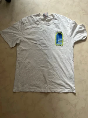 Vit Casa Blanca t-shirt med tryck - Säljer en vit t-shirt från Casa Blanca med ett färgglatt tryck på bröstet i blått och gult. Klassisk rund hals och korta ärmar. Perfekt för dig som gillar stilrena plagg med en twist. Storlek XL går att diskutera pris