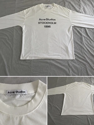 Vit långärmad Acne Studios t-shirt - Vit långärmad t-shirt från Acne Studios med svart text 'Acne Studios STOCKHOLM 1996' på bröstet. Klassisk rund halsringning och rak passform. Tillverkad i mjuk bomull som känns skön mot huden. Perfekt för en clean och stilren look.