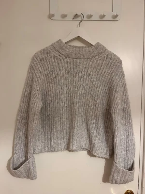Ljusgrå stickad tröja med polokrage - Mysig ljusgrå stickad tröja med bred ribbstickning och hög pololiknande krage. Tröjan har långa ärmar med uppvikta muddar och en något boxig, avslappnad passform. Perfekt för kalla dagar och enkel att matcha med jeans eller kjol.