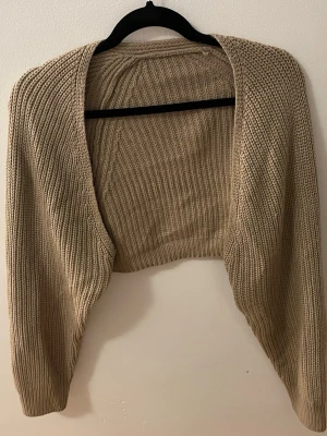 Beige stickad bolero - Säljer en beige stickad bolero med öppen front och långa ärmar. Plagget har en grovstickad struktur och är perfekt att slänga över en topp för extra värme. Materialet känns mjukt och stretchigt, vilket gör den bekväm att bära. 