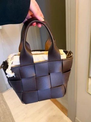 Brun flätad handväska i läder - Snygg brun handväska i flätad läderdesign med två korta handtag och en beige tygpåse inuti som stängs med dragsko. Väskan har en kompakt och boxig form, perfekt för att bära det viktigaste. Passar dig som gillar trendiga accessoarer.
