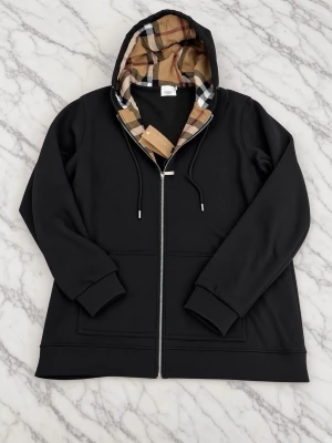 Svart hoodie med Burberry-rutor - Snygg svart hoodie från Burberry med dragkedja och klassiskt rutigt foder i huvan. Hoodien har dragsko i huvan, ribbade muddar och diskret Burberry-logga broderad på bröstet. Perfekt för dig som vill ha en stilren och exklusiv look.