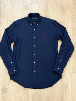 Mörkblå linneskjorta från Polo Ralph Lauren - Klassisk mörkblå skjorta i linne från Polo Ralph Lauren med vit knappstängning och röd broderad logga på bröstet. Skjortan har lång ärm, button-down krage och en avslappnad passform. Perfekt till jeans eller chinos.