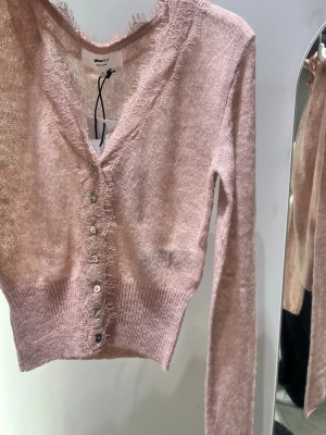 Ljusrosa cardigan med spetskant - Supersöt ljusrosa cardigan med långa ärmar och v-ringning. Tröjan har dekorativa spetsdetaljer längs kanten och knäpps med små pärlemorknappar framtill. Materialet är mjukt och lätt stickat, perfekt för lager på lager.