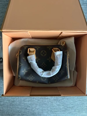 Brun handväska från Louis Vuitton speedy - Snygg brun handväska från Louis Vuitton med axelrem och guldfärgade detaljer. Kommer i originalförpackning och med LV-logga. Perfekt accessoar för dig som gillar exklusiv och stilren design. Louis Vuitton Speedy bag. Helt oanvänd 
